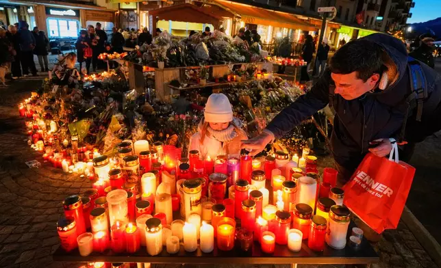 FILE - A child watches as an adult places a candle outside the sealed off Le Constellation bar in Crans-Montana, Swiss Alps, Switzerland, Saturday, Jan. 3, 2026, where a devastating fire left dead and injured during the New Year's celebrations. (AP Photo/Baz Ratner, File)