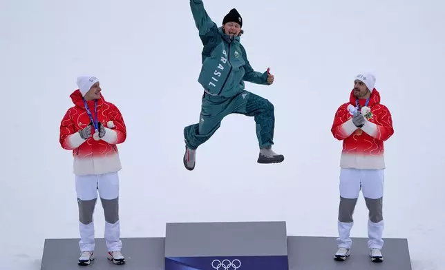Brazil's Lucas Pinheiro Braathen, center, winner of an alpine ski, men's giant slalom race, jumps in celebration on the podium flanked by second placed Switzerland's Marco Odermatt, left, and third placed Switzerland's Loic Meillard, at the 2026 Winter Olympics, in Bormio, Italy, Saturday, Feb. 14, 2026. (AP Photo/John Locher)