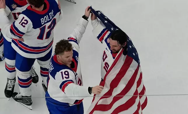 United States' Matthew Tkachuk (19) helps put an American flag around goalie Connor Hellebuyck after the United States beat Canada in overtime to win the men's gold medal hockey game at the 2026 Winter Olympics, in Milan, Italy, Sunday, Feb. 22, 2026. (AP Photo/David J. Phillip)