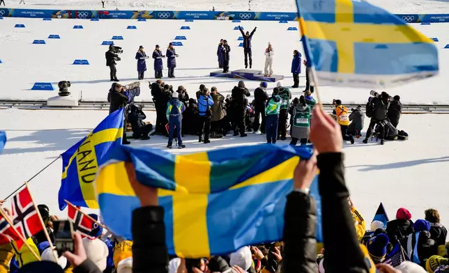 Sweden fans wave flags as Frida Karlsson, of Sweden, jumps on the podium celebrating after winning the gold medal in the cross country skiing women's 10km interval start free at the 2026 Winter Olympics, in Tesero, Italy, Thursday, Feb. 12, 2026. (AP Photo/Evgeniy Maloletka)
