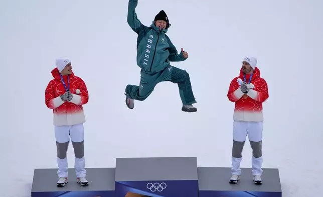 Brazil's Lucas Pinheiro Braathen, center, winner of an alpine ski, men's giant slalom race, jumps in celebration on the podium flanked by second placed Switzerland's Marco Odermatt, left, and third placed Switzerland's Loic Meillard, at the 2026 Winter Olympics, in Bormio, Italy, Saturday, Feb. 14, 2026. (AP Photo/John Locher)