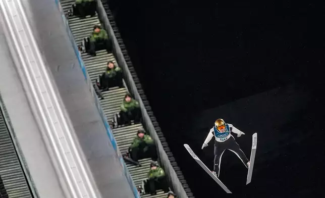 Italian Army soldiers on the steps of the ramp watch Domen Prevc, of Slovenia, soaring through the air during his first round jump of the ski jumping men's large hill individual at the 2026 Winter Olympics, in Predazzo, Italy, Saturday, Feb. 14, 2026. (AP Photo/Matthias Schrader)