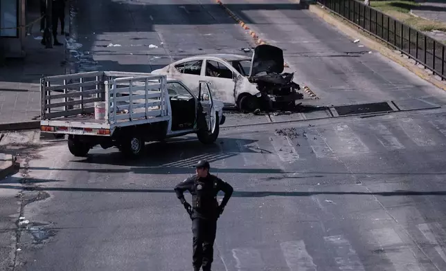 A police officer stands guard by a charred vehicle after it was set on fire, on a road in Guadalajara, Jalisco state, Mexico, Sunday, Feb. 22, 2026, after the death of the leader of the Jalisco New Generation Cartel, Nemesio Rubén Oseguera Cervantes, known as "El Mencho." (AP Photo/Alejandra Leyva)