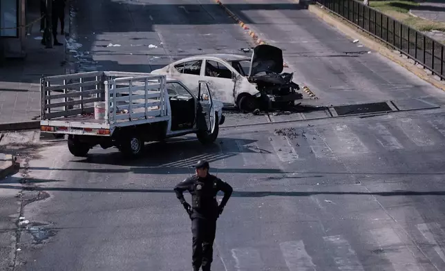 A police officer stands guard by a charred vehicle after it was set on fire, on a road in Guadalajara, Jalisco state, Mexico, Sunday, Feb. 22, 2026, after the death of the leader of the Jalisco New Generation Cartel, "El Mencho." (AP Photo/Alejandra Leyva)