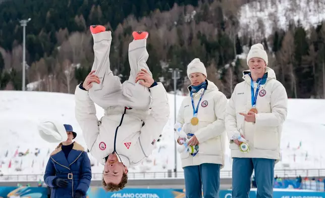 Silver medalist Ben Ogden, of the United States, does a back flip from the podium while gold medalist Johannes Hoesflot Klaebo, of Norway, and bronze medalist Oskar Opstad Vike, also of Norway, right, look on after the cross-country skiing men's sprint classic at the 2026 Winter Olympics, in Tesero, Italy, Tuesday, Feb. 10, 2026. (AP Photo/Evgeniy Maloletka)