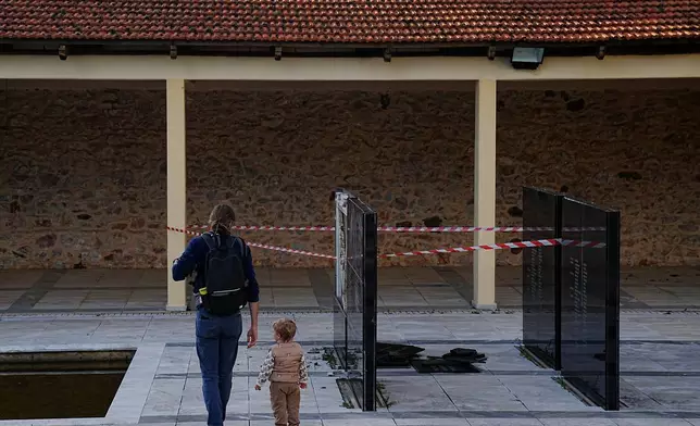 A man with a child walk past a vandalised memorial plaque at the Kaisariani Shooting Range in the Kaisariani suburb of Athens, where part of a black marble monument bearing the names of 200 Greek communist political prisoners executed by Nazi forces in 1944 was smashed, Monday, Feb. 16, 2026. (AP Photo/Petros Giannakouris)