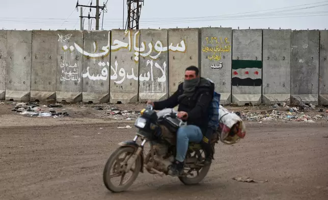 A man rides his motorcycle past the wall of a U.S. military base bearing graffiti in Arabic that reads, "Syria is more beautiful, no Assad, no SDF," and in yellow, "Al-Shaddadi without SDF terrorism," near the village of Al-Shaddadi, al-Hassakeh province, eastern Syria, Monday, Feb. 2, 2026. (AP Photo/Ghaith Alsayed)