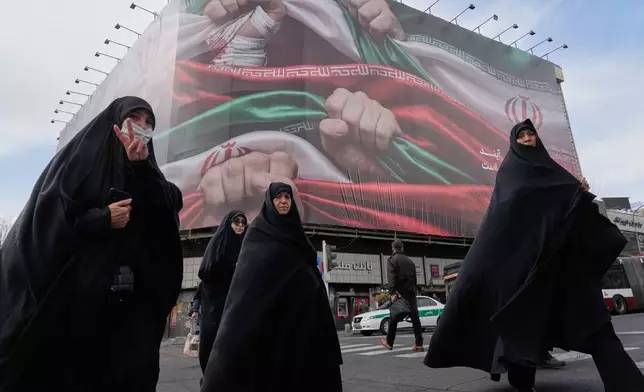 FILE - Women, one flashing a victory hand gesture, cross a street under a huge banner showing hands firmly holding Iranian national flags as a sign of patriotism, in Tehran, Iran, Jan. 14, 2026. (AP Photo/Vahid Salemi, File)