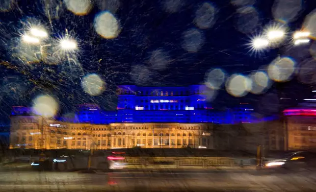 A passing car splashes water from a puddle onto a camera lens as photos are taken of the Palace of Parliament illuminated in the colors of the Ukrainian flag, to mark the fourth anniversary of the Russian invasion of Ukraine, in Bucharest, Romania, Tuesday, Feb. 24, 2026. (AP Photo/Andreea Alexandru)