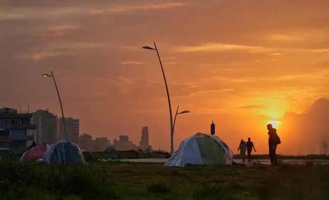 People walk past tents sheltering people displaced by Israeli airstrikes at a public space along the Beirut waterfront at sunset in Beirut, Lebanon, Sunday, March 15, 2026. (AP Photo/Hassan Ammar)