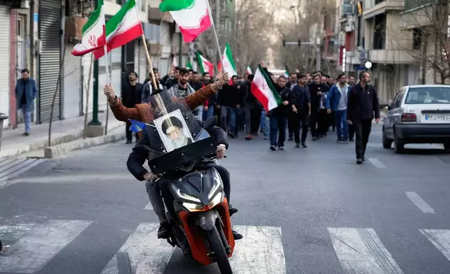 Two men on a motorcycle decorated with a picture of the late Iranian Supreme Leader Ayatollah Ali Khamenei ride ahead a group of government supporters near Khamenei's residency in Tehran, Iran, Sunday, March 1, 2026, in the aftermath of his confirmed death in U.S. and Israeli strikes. (AP Photo/Vahid Salemi)