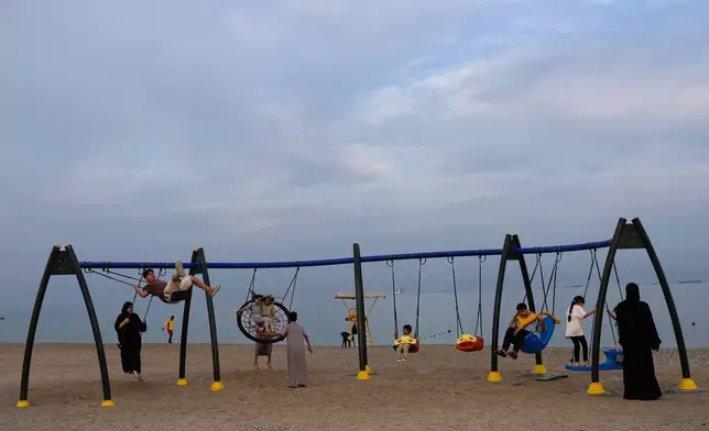 Children and adults play on swings on the beach as oil tankers and cargo ships line up in the Strait of Hormuz near Khor Fakkan, United Arab Emirates, Wednesday, March 11, 2026. (AP Photo/Altaf Qadri)