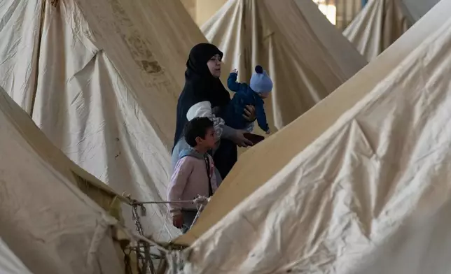 A displaced woman holds a child as another stands beside her between rows of tents at the Camille Chamoun Sports City Stadium, which has been turned into a shelter for people displaced by Israeli airstrikes in southern Lebanon and Dahiyeh, Beirut's southern suburbs, in Beirut, Lebanon, Tuesday, March 10, 2026. (AP Photo/Hassan Ammar)