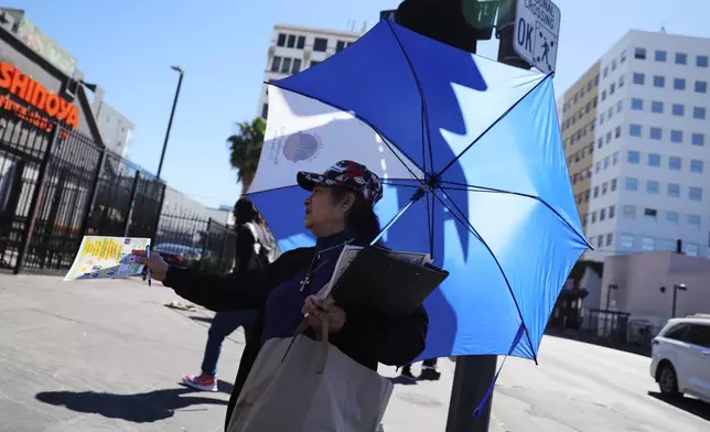 A woman covers herself from the sun while handing out fliers during an unseasonably hot day at MacArthur Park on Thursday, March 12, 2026, in Los Angeles. (AP Photo/Ryan Sun)