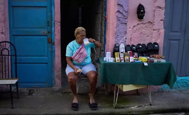 A vendor having breakfast sits by her table holding various products, from cigarettes to sandals, in Havana, Cuba, early Friday, March 13, 2026. (AP Photo/Ramon Espinosa)