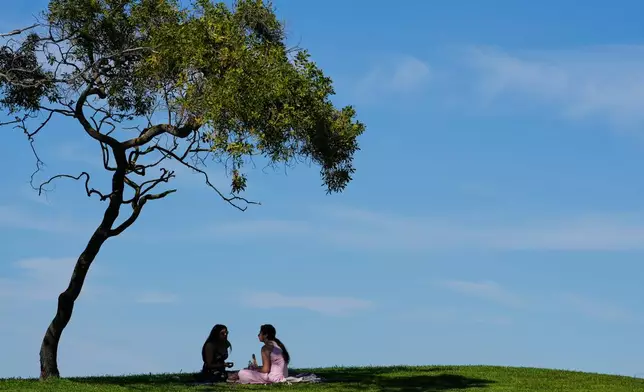 Two women sit in the shade under a tree while visiting Shoreline Park in Mountain View, Calif., Monday, March 16, 2026. (AP Photo/Godofredo A. Vásquez)
