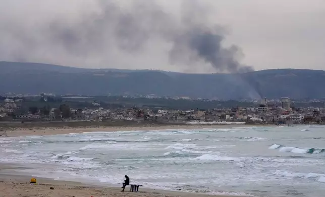 A man checks his mobile phone on a public beach, as smoke, background, rises from Israeli artillery shells on Qlaileh village, as it s seen from Tyre city, south Lebanon, Saturday, March 28, 2026. (AP Photo/Hussein Malla)