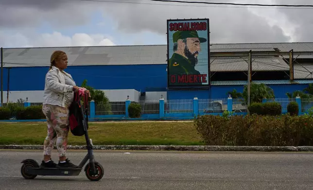 A woman rides an electric scooter past a factory displaying an image depicting the late Cuban leader Fidel Castro, bearing the words "Socialism or Death", in Havana, Cuba, Thursday, March 19, 2026. (AP Photo/Ramon Espinosa)