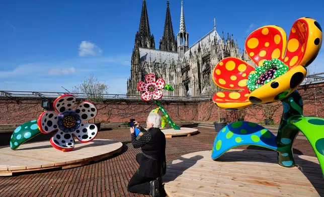A woman takes pictures of the artwork 'Flowers that speak all about my heart given to the sky' in front of the Cologne Cathedral during a preview of the new major exhibition of legendary Japanese artist Yayoi Kusama at the Museum Ludwig in Cologne, Thursday, March 12, 2026. (AP Photo/Martin Meissner)