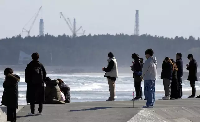 People observe a moment of silence at 2:46 p.m., the moment the earthquake struck with a backdrop of Fukushima Daiichi nuclear power plant in Namie, Fukushima prefecture, northern Japan as the country marked the 15th anniversary of the massive earthquake, tsunami and nuclear disaster. (Kyodo News via AP)