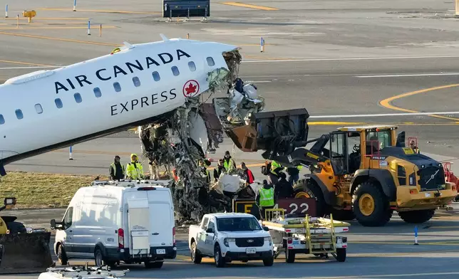 Aircraft maintenance workers position a front loader under debris hanging from the wreckage of an Air Canada Express jet, Tuesday, March 24, 2026, just off the runway where it had collided with a Port Authority fire truck Sunday night at LaGuardia Airport in New York. (AP Photo/Yuki Iwamura)