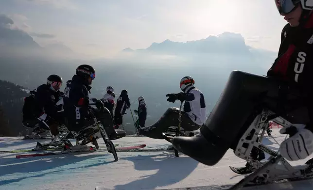 A Swiss athlete, right, rides by while athletes from the United States check the track before the start of the day's alpine skiing competitions at the 2026 Winter Paralympics, in Cortina d'Ampezzo, Italy, Tuesday, March 10, 2026. (AP Photo/Evgeniy Maloletka)