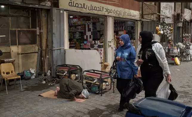Women walk past a man praying on the sidewalk in downtown of Baghdad, Iraq, Wednesday, March 25, 2026. (AP Photo/Leo Correa)