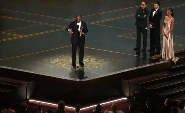 Ryan Coogler accepts the award for writing (original screenplay) for "Sinners" during the Oscars on Sunday, March 15, 2026, at the Dolby Theatre in Los Angeles. Robert Downey Jr., second right, and Chris Evans looks on from right.(AP Photo/Chris Pizzello)