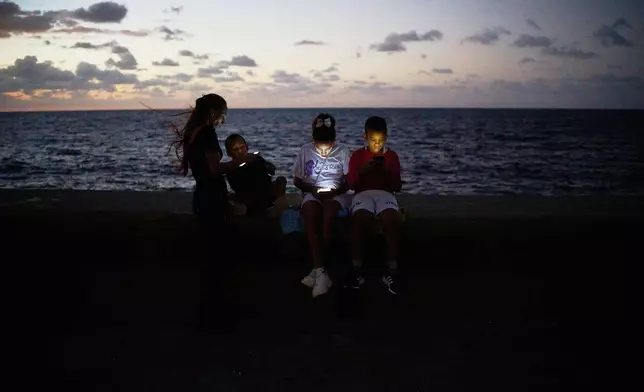 Children look at their phones while sitting on the Malecón wall during a blackout in Havana, Cuba, Saturday, March 21, 2026. (AP Photo/Ramon Espinosa)