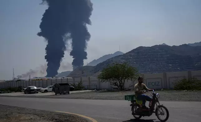 A man rides a bike as fires and plumes of smoke rise after debris from an intercepted Iranian drone struck an oil facility, according to authorities, in Fujairah, United Arab Emirates, Saturday, March 14, 2026. (AP Photo/Altaf Qadri)