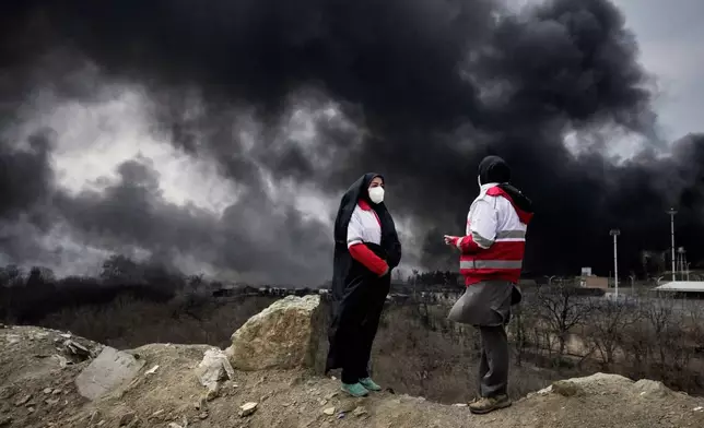FILE - Two women from the Iranian Red Crescent Society stand as a thick plume of smoke from a U.S.-Israeli strike on an oil storage facility late Saturday rises into the sky in Tehran, Iran, Sunday, March 8, 2026. (AP Photo/Vahid Salemi, File)
