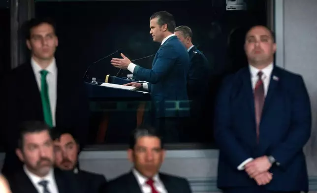 Staff members watch as Defense Secretary Pete Hegseth, reflected in a window, speaks during a press briefing at the Pentagon, Monday, March 2, 2026, in Washington. (AP Photo/Mark Schiefelbein)