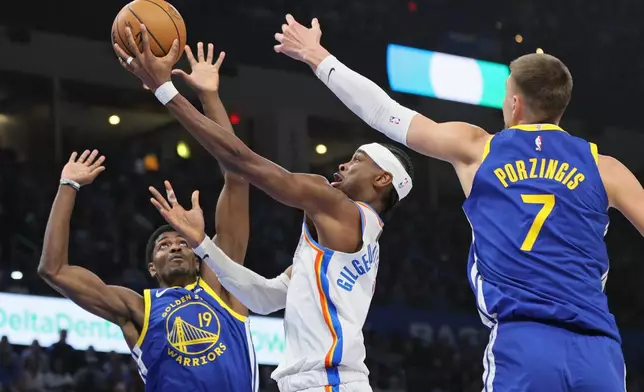 Oklahoma City Thunder guard Shai Gilgeous-Alexander, center, takes the ball to the basket between Golden State Warriors guard Nate Williams (19) and center Kristaps Porzingis (7) during the first half of an NBA basketball game Saturday, March 7, 2026, in Oklahoma City. (AP Photo/Nate Billings)