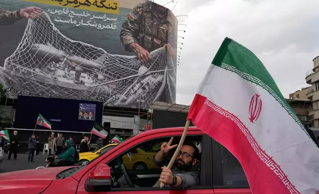 A man flashes a victory sign as he carries an Iranian flag in front of an anti-U.S. billboard depicting the American aircrafts into the Iranian armed forces fishing net with signs that read in Farsi: "The Strait of Hormuz will remain closed, The entire Persian Gulf is our hunting ground," at the Eqelab-e-Eslami, or Islamic Revolution Square in downtown Tehran, Iran, Monday, April 13, 2026. (AP Photo/Vahid Salemi)