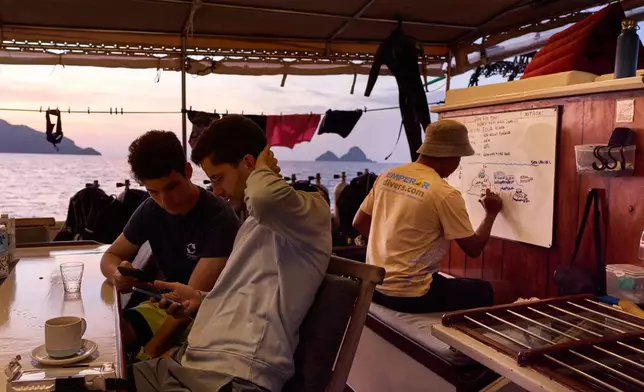 Scuba divers Alejandro Sarró, left, and Manel Sangrà check their phones as dive guide Kristanto Umbu Kudu prepares a briefing aboard the Raja Laut dive boat in Raja Ampat, Indonesia, Tuesday, March 3, 2026. (AP Photo/Claudia Rosel)