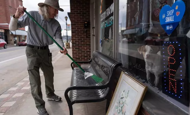 Philip Moulton sweeps the bench outside his Ravenswood Arts gallery where his dog JoJo peers through window, Friday, March 13, 2026, in Ravenswood W.Va., where surging electricity costs have forced some stores out of business. (AP Photo/Carolyn Kaster)