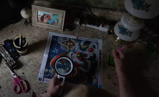 Rebecca Michalski works on her diamond painting kit by window light, in an effort to save money on her electricity bill, at her home in Rainelle, W.Va., Saturday, March 21, 2026, next to a photo of her daughter Sabrina Nicole Adkins, who passed away last year and whose funeral costs were difficult to afford. (AP Photo/Carolyn Kaster)