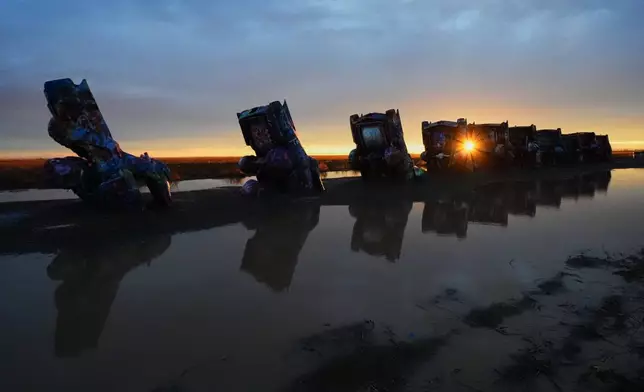 A puddle from a midday storm surrounds vehicles as the setting sun peeks through at Cadillac Ranch, a roadside attraction along Route 66 in Amarillo, Texas, Thursday, Nov. 20, 2025. (AP Photo/Julio Cortez)