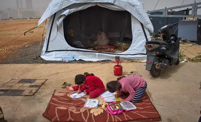 Tamara and her sister Amal color pictures on the floor as their parents, Sara and Ahmed, who fled their village of Khiyam in southern Lebanon due to Israeli bombardment, sit inside a tent used as a shelter in Beirut, Lebanon, Friday, April 3, 2026. (AP Photo/Emilio Morenatti)