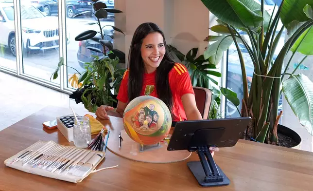 Artist Lili Cantero pulls up an image she is working from on a screen as she paints a soccer ball commemorating Spain's 2010 World Cup victory as part of the launch of the Wynwood United Soccer Ball Art Trail, which will showcase hand-painted soccer balls honoring historic World Cup championships inside local businesses, April 8, 2026, at Rodilla cafe in the Wynwood district of Miami. (AP Photo/Rebecca Blackwell)