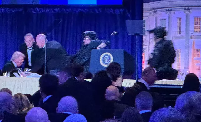 President Donald Trump, left, is seated on stage as U.S. Secret Service agents respond to take him from the ballroom after a shooting incident outside the ballroom during the White House Correspondents Dinner, Saturday, April 25, 2026, in Washington. (AP Photo/Mark Schiefelbein)