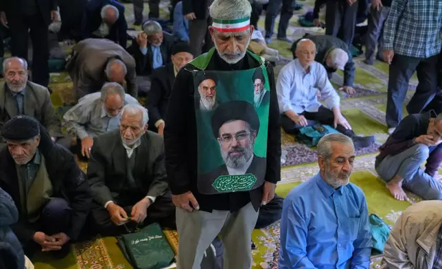An Iranian worshipper wears a banner showing portraits of the Supreme Leader Ayatollah Mojtaba Khamenei, bottom, late Supreme Leader Ayatollah Ali Khamenei, top right, and the late revolutionary founder Ayatollah Khomeini, during Friday prayers ceremony at the Tehran University campus, in Tehran, Iran, Friday, April 24, 2026. (AP Photo/Vahid Salemi)