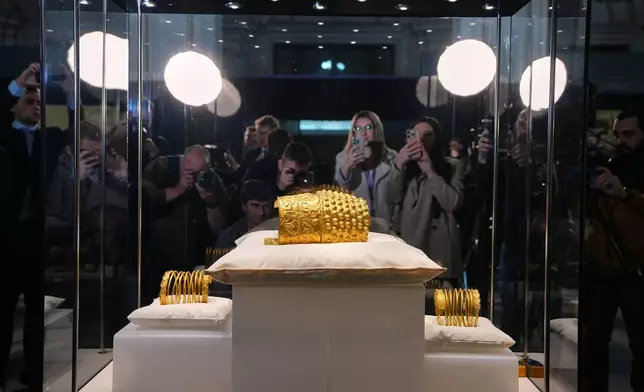 Journalists gather around a glass enclosure with the Dacian gold items, a 2,500-year-old helmet and wristbands, stolen from a museum in the Netherlands and then recovered by Dutch authorities, after they were presented upon being returned at the National Museum of Romanian History, in Bucharest, Romania, Tuesday, April 21, 2026. (AP Photo/Andreea Alexandru)