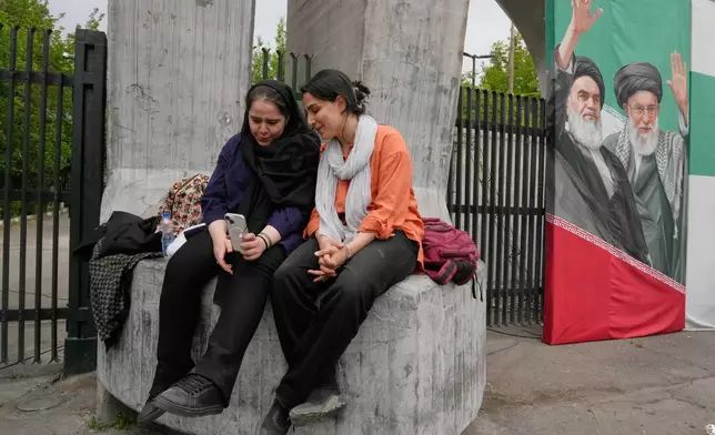Women share a moment as they look at a smartphone at the main gate of the Tehran University as a banner shows portraits of the late Supreme Leader Ayatollah Ali Khamenei, right, and the late revolutionary founder Ayatollah Khomeini in Tehran, Iran, Sunday, April 19, 2026. (AP Photo/Vahid Salemi)