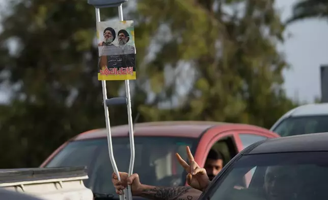 A displaced man flashes victory sign and holds a crutch with a poster showing late Hezbollah leaders Sayyed Hassan Nasrallah and Hashem Safieddine, with writing that reads "on the pledge, we remain" as he returns to his village following a ceasefire between Hezbollah and Israel, in Qasmiyeh near the city of Tyre, south Lebanon, Friday, April 17, 2026. (AP Photo/Mohammed Zaatari)
