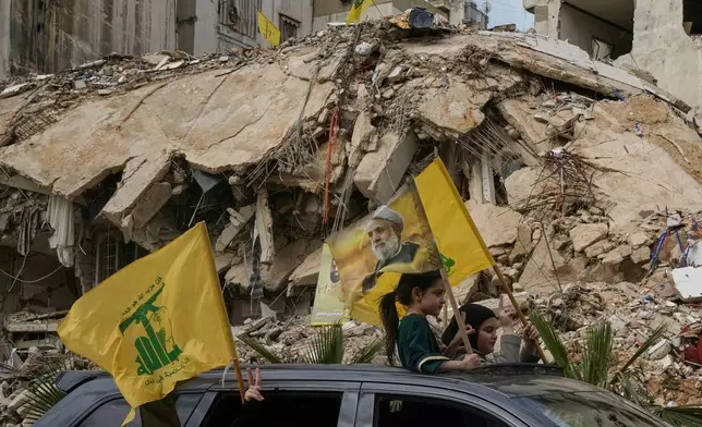Displaced residents wave Hezbollah flags, including one bearing a picture of its leader, Naim Qassem, as they pass rubble of destroyed buildings in Dahiyeh, Beirut's southern suburbs, Lebanon, Friday, April 17, 2026, following a ceasefire between Israel and Hezbollah. (AP Photo/Bilal Hussein)