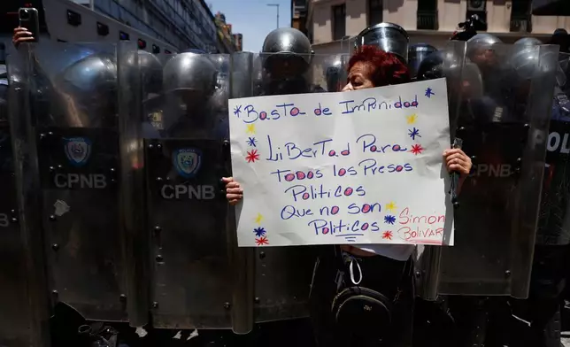 A protester holds up a sign demanding freedom for all political prisoners as she stands in front of a cordon of Bolivarian National Police, during a march demanding higher salaries, pensions and benefits, in Caracas, Venezuela, Thursday, April 9, 2026. (AP Photo/Pedro Mattey)