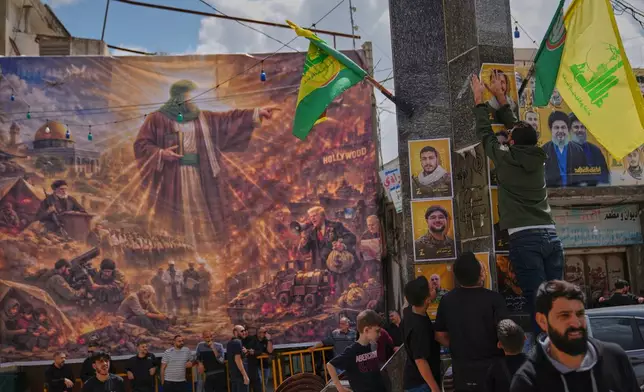 Hezbollah supporters hang portraits of fighters killed in Israeli airstrikes beside a large billboard depicting the war in the Middle East following U.S.-Israeli strikes on Iran on Feb. 28, ahead of a funeral procession in the southern village of Kfar Sir, Lebanon, Tuesday, April 21, 2026. (AP Photo/Hassan Ammar)