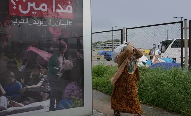 FILE — A displaced woman who fled Israeli airstrikes in southern Lebanon, carries her belonging as she moves to a better spot to shelter from the rain, past an Arabic anti-war poster that reads, "Sacrificing for whom? Lebanon does not need war," in Beirut, Saturday, March 21, 2026. (AP Photo/Hussein Malla, File)