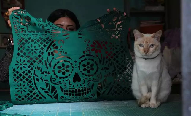 Cande Martinez prepares soccer jerseys inspired by papel picado and traditional folk art, alongside her cat Nube in Cuautitlán Izcalli, Mexico, Monday, May 25, 2026. (AP Photo/Marco Ugarte)
